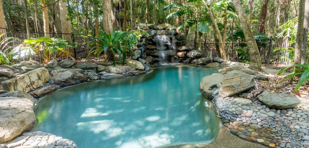 Tranquil natural pool with a waterfall, surrounded by rocks and lush greenery in a forest setting. Sunlight filters through the trees, creating a serene atmosphere.