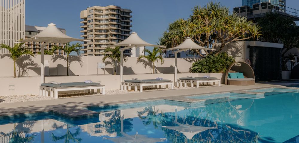 A tranquil outdoor pool with clear blue water reflects nearby white canopies and palm trees. Modern high-rise buildings sit under a clear blue sky.