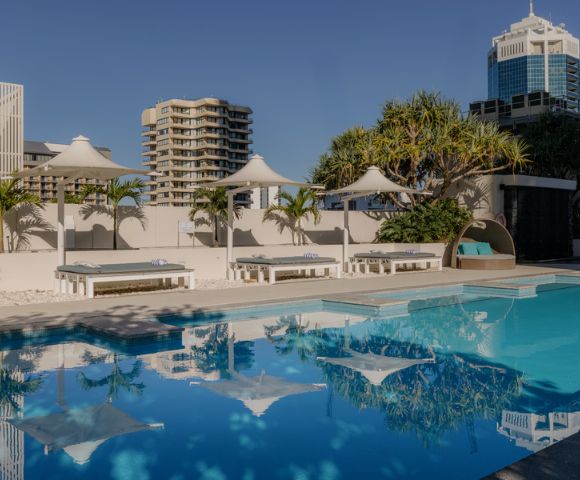 A tranquil outdoor pool with clear blue water reflects nearby white canopies and palm trees. Modern high-rise buildings sit under a clear blue sky.
