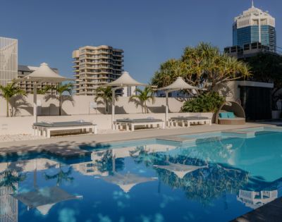 A tranquil outdoor pool with clear blue water reflects nearby white canopies and palm trees. Modern high-rise buildings sit under a clear blue sky.