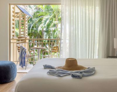 A serene bedroom with a sun hat and scarf on the bed. Open balcony doors reveal lush greenery, creating a fresh and tranquil ambiance.