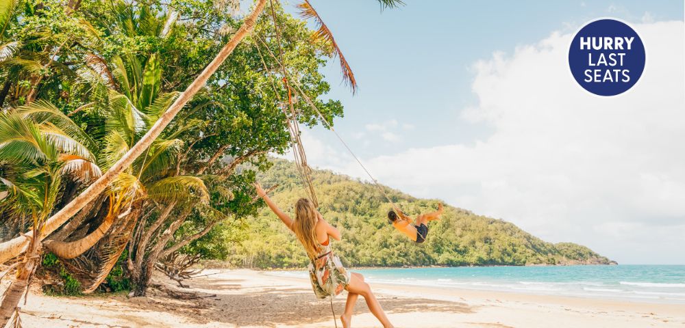 A woman swings on a beach surrounded by palm trees, with a scenic view of the ocean and mountains, highlighted by a 