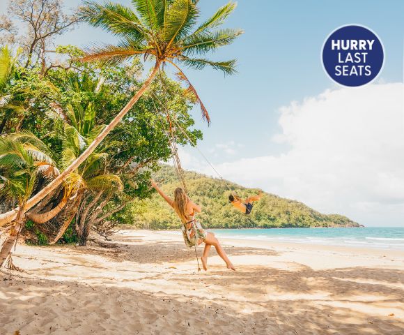 A woman swings on a beach surrounded by palm trees, with a scenic view of the ocean and mountains, highlighted by a 