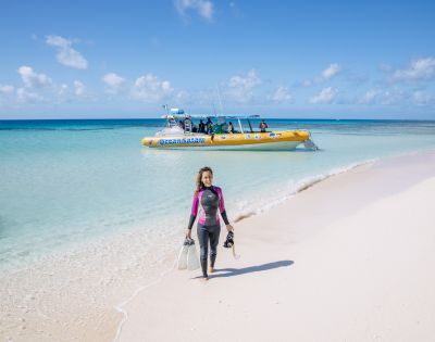 A woman in a wetsuit walks on a white sandy beach carrying snorkeling gear, with a yellow boat and turquoise sea in the background under a clear sky.