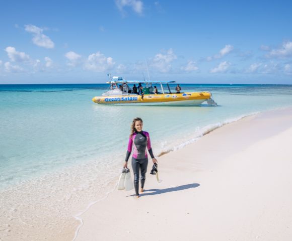 A woman in a wetsuit walks on a white sandy beach carrying snorkeling gear, with a yellow boat and turquoise sea in the background under a clear sky.