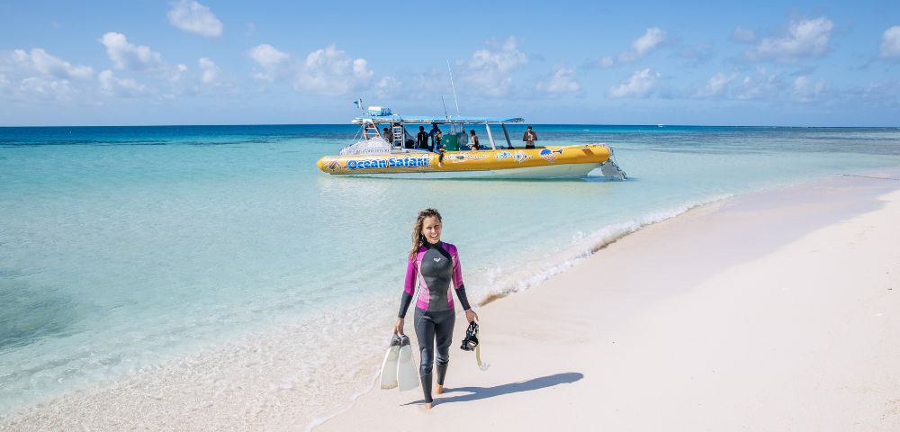 A woman in a wetsuit walks on a white sandy beach carrying snorkeling gear, with a yellow boat and turquoise sea in the background under a clear sky.
