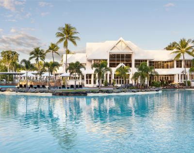 A serene resort with a modern white building, surrounded by palm trees and a turquoise lagoon pool under a clear sky.