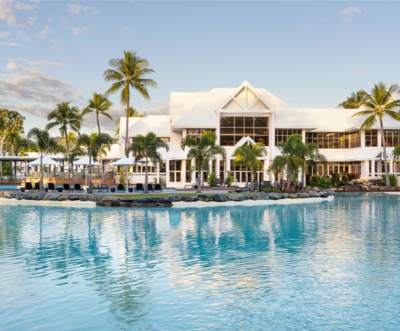 A serene resort with a modern white building, surrounded by palm trees and a turquoise lagoon pool under a clear sky.