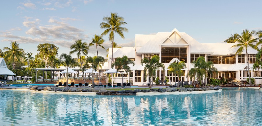 A serene resort with a modern white building, surrounded by palm trees and a turquoise lagoon pool under a clear sky.