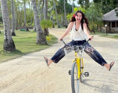 Young woman with dark curly hair, wearing a white top and patterned pants, excitedly rides a yellow bicycle on a sandy path with legs spread wide, eyes wide, and mouth open, near palm trees.