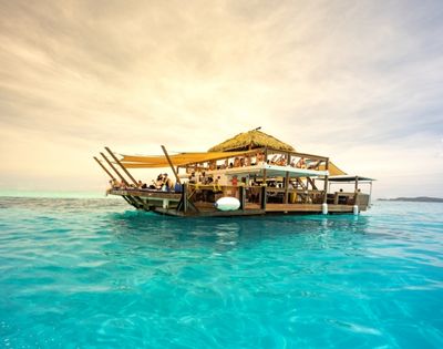 A lively floating bar with a thatched roof, surrounded by turquoise waters, filled with beachgoers enjoying drinks and sun.