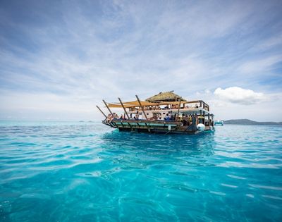 A lively floating restaurant with a thatched roof, surrounded by crystal-clear turquoise waters under a bright sky.