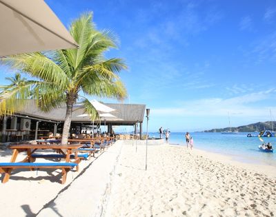 Beachfront restaurant with picnic tables, palm trees, and umbrellas, with a white sand beach and clear blue water visible in the background, people walking on the beach and kayaking.