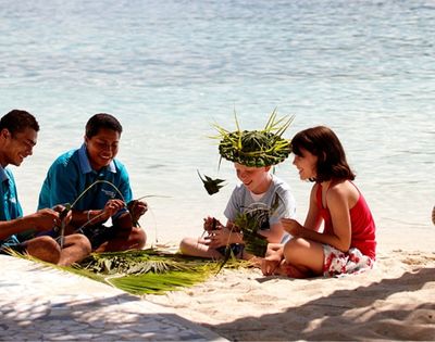 Four people sitting on a sandy beach with the ocean in the background. Two men in blue shirts, and two children are making crafts from green leaves, with one child wearing a leaf hat. A palm tree trunk is on the right side of the image.