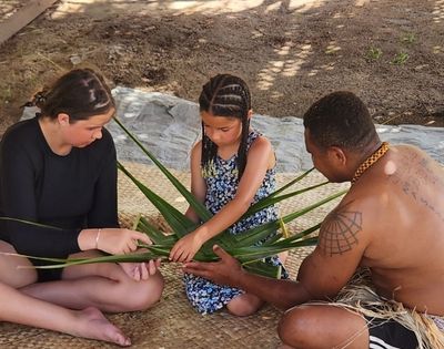 Three people weaving palm fronds together while sitting on a woven mat. The person on the right, a man, is shirtless with a thick gold necklace and tattoos on his back. Two young girls are on the left and center wearing a black shirt and floral sundress, respectively. They're all focused on manipulating the leaves.