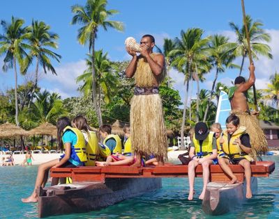 A Polynesian man in traditional attire blows into a conch shell while standing on a watercraft, surrounded by children wearing life vests, with a tropical beach scene in the background under a bright blue sky.