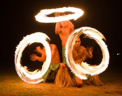 Three shirtless men performing a fire dance at night, each spinning flaming hoops around themselves and in the air, the flames creating light streaks.