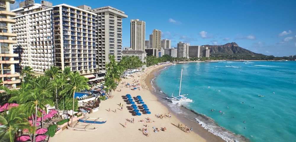 A scenic view of a sandy beach with blue umbrellas, hotels, palm trees, and a calm turquoise ocean, featuring Diamond Head in the background.