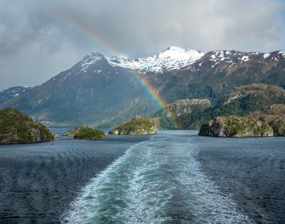 A vibrant rainbow arcs over a snowy mountain and rocky islands, reflected in the serene blue water below. The scene is tranquil and picturesque.