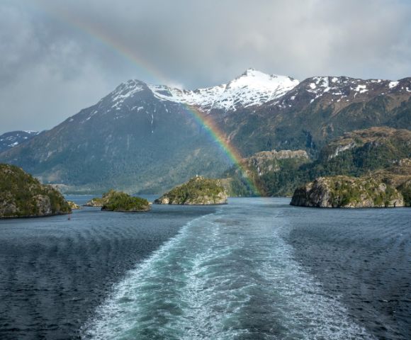 A vibrant rainbow arcs over a snowy mountain and rocky islands, reflected in the serene blue water below. The scene is tranquil and picturesque.