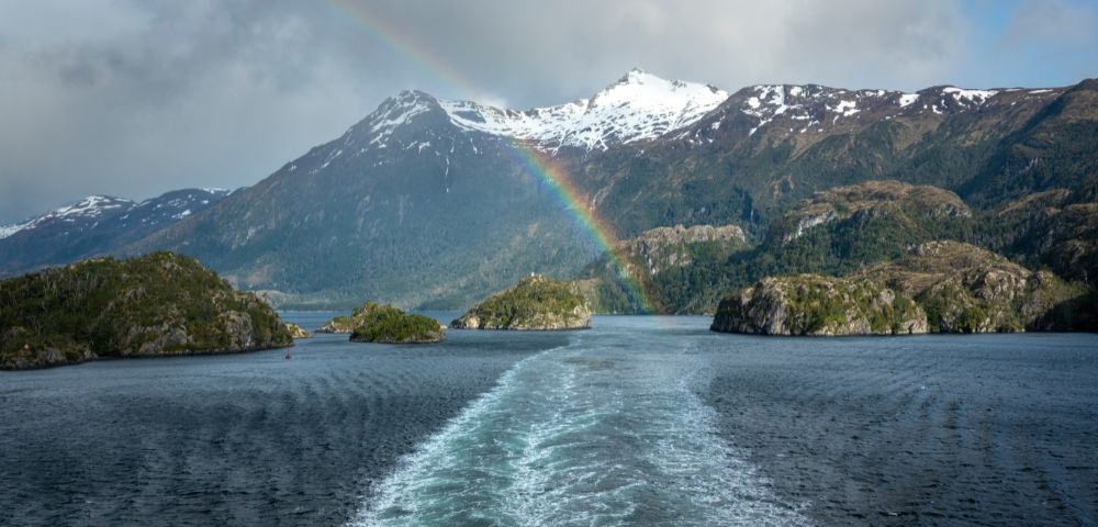 A vibrant rainbow arcs over a snowy mountain and rocky islands, reflected in the serene blue water below. The scene is tranquil and picturesque.