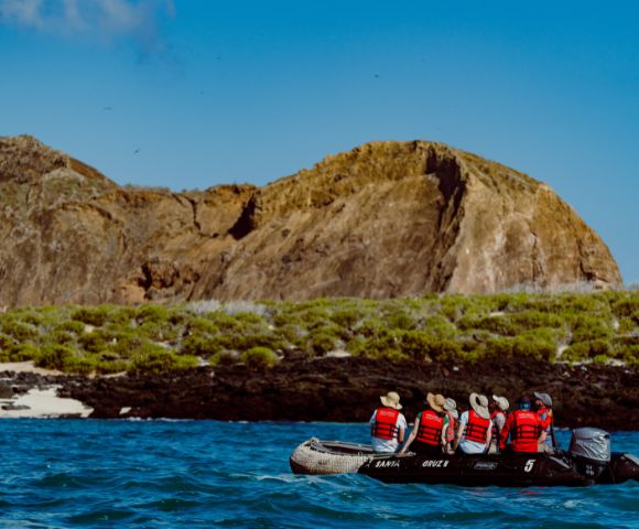 A small boat with six people in red life jackets approaches a rocky island with green vegetation under a clear blue sky, conveying adventure and exploration.