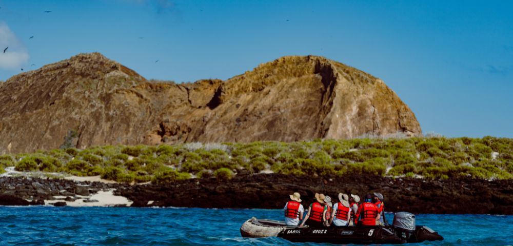 A small boat with six people in red life jackets approaches a rocky island with green vegetation under a clear blue sky, conveying adventure and exploration.