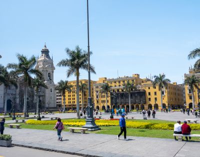 A vibrant city square on a sunny day with people walking and relaxing. Surrounded by palm trees, historic architecture, and a yellow colonial building.