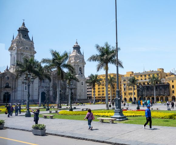 A vibrant city square on a sunny day with people walking and relaxing. Surrounded by palm trees, historic architecture, and a yellow colonial building.