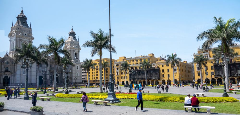 A vibrant city square on a sunny day with people walking and relaxing. Surrounded by palm trees, historic architecture, and a yellow colonial building.
