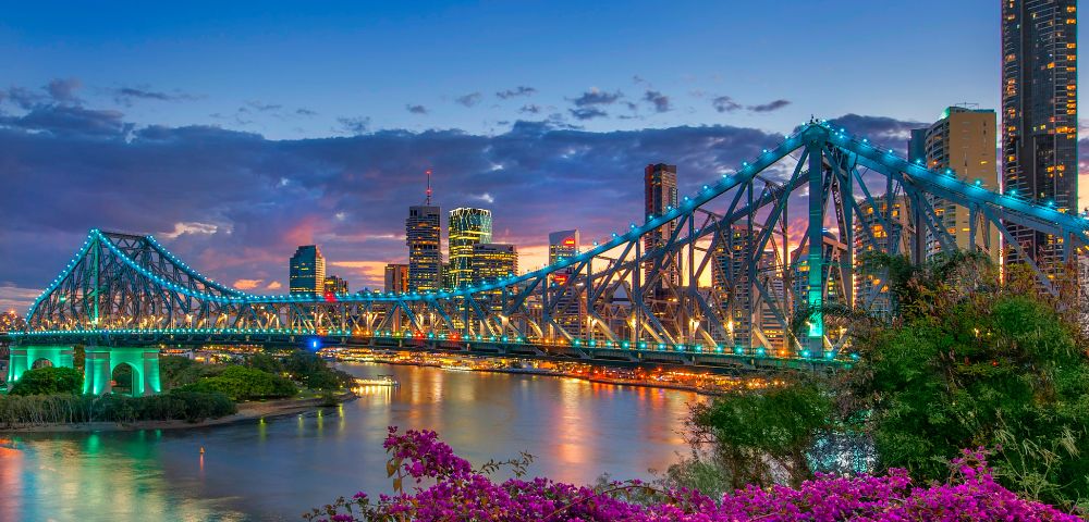 Brisbane's Story Bridge illuminated at twilight, with vibrant city skyline and blooming purple bougainvillea in the foreground.