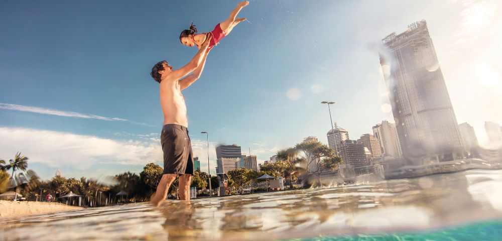 A joyful father lifts his laughing daughter high above the water on a sunny day, with a city skyline in the background.