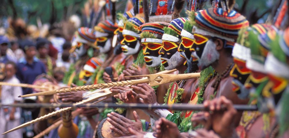 A vibrant line of dancers with painted faces and colorful headdresses playing traditional instruments at a cultural festival.