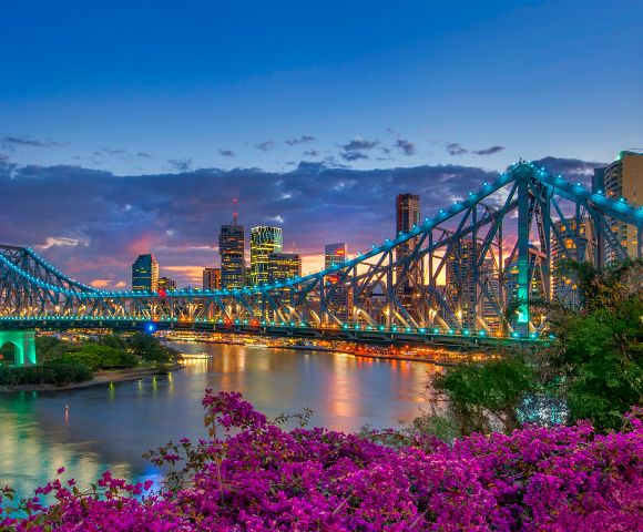 Brisbane's Story Bridge illuminated at twilight, with vibrant city skyline and blooming purple bougainvillea in the foreground.