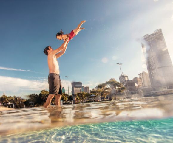 A joyful father lifts his laughing daughter high above the water on a sunny day, with a city skyline in the background.