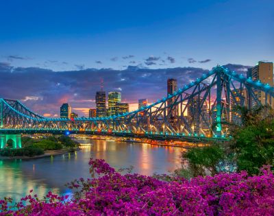 Brisbane's Story Bridge illuminated at twilight, with vibrant city skyline and blooming purple bougainvillea in the foreground.