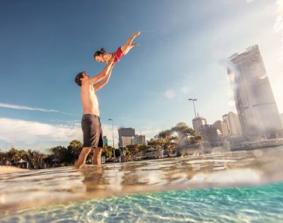 A joyful father lifts his laughing daughter high above the water on a sunny day, with a city skyline in the background.