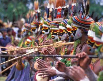 A vibrant line of dancers with painted faces and colorful headdresses playing traditional instruments at a cultural festival.