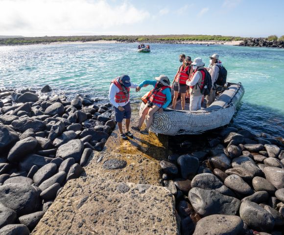 A group of six people in life jackets disembark from a small boat on rocky terrain by a turquoise sea in a sunny coastal setting.