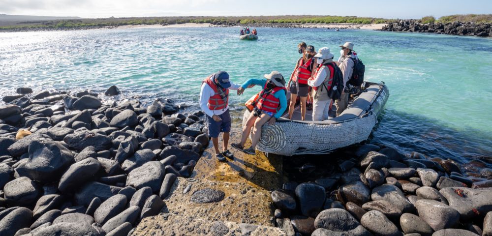 A group of six people in life jackets disembark from a small boat on rocky terrain by a turquoise sea in a sunny coastal setting.