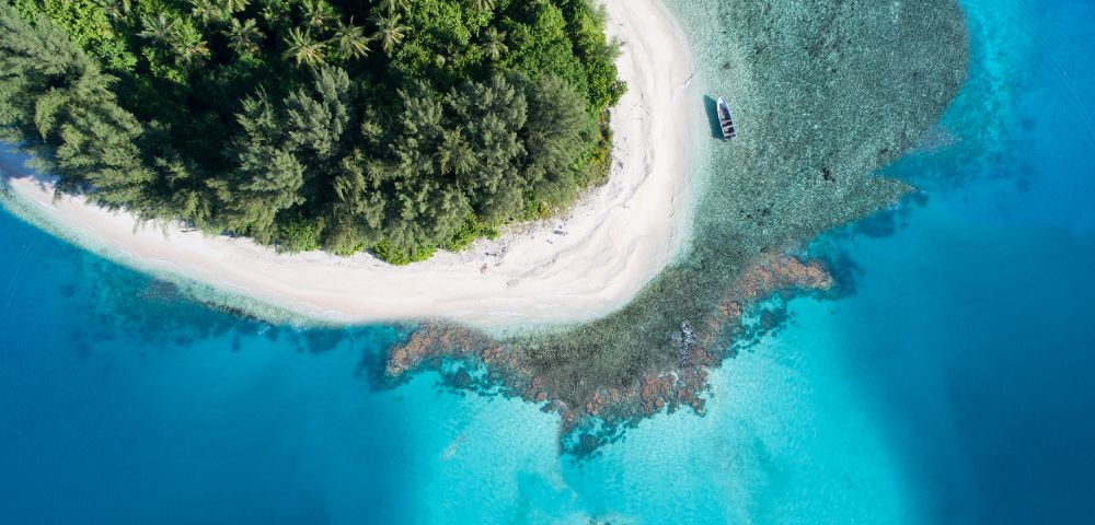 Aerial view of a tropical island with a white sand beach, lush green vegetation, and turquoise waters. A small boat is moored near the shore. The water transitions from clear shallows over coral reefs to deep blue ocean.