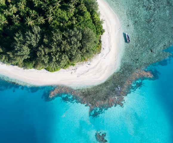 Aerial view of a tropical island with a white sand beach, lush green vegetation, and turquoise waters. A small boat is moored near the shore. The water transitions from clear shallows over coral reefs to deep blue ocean.