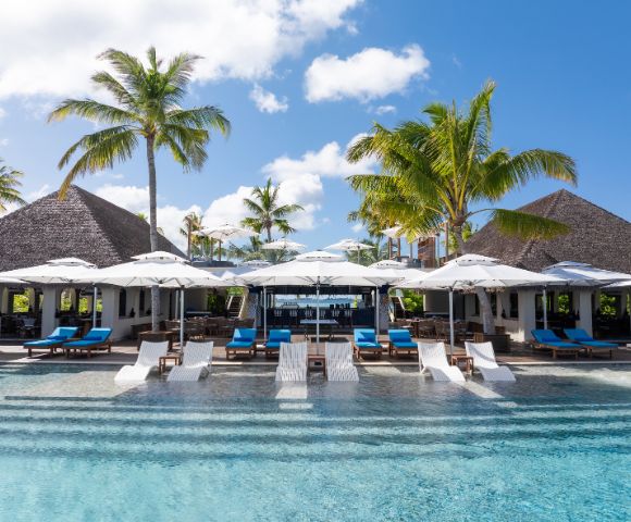 Tropical resort pool with lounge chairs, umbrellas, and palm trees in a serene setting, featuring a restaurant with a thatched roof.