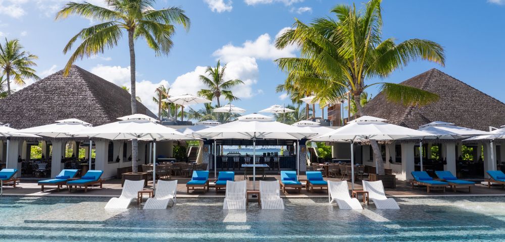 Tropical resort pool with lounge chairs, umbrellas, and palm trees in a serene setting, featuring a restaurant with a thatched roof.