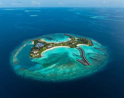 Aerial view of a tropical island resort surrounded by turquoise waters and coral reefs, featuring villas and lush greenery.