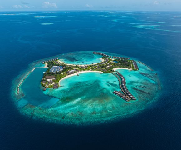 Aerial view of a tropical island resort surrounded by turquoise waters and coral reefs, featuring villas and lush greenery.