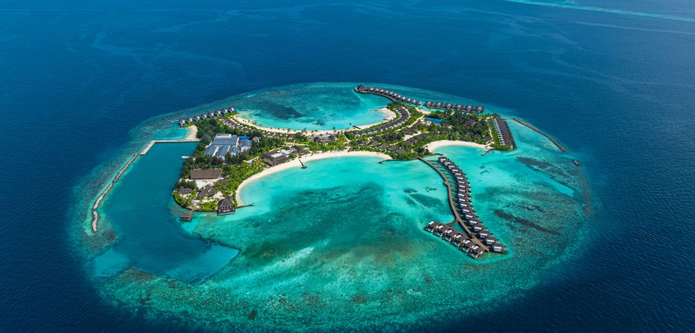 Aerial view of a tropical island resort surrounded by turquoise waters and coral reefs, featuring villas and lush greenery.