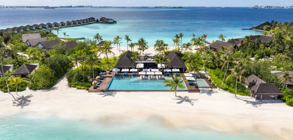 Aerial view of a tropical resort with a pool, sun loungers, and palm trees, adjacent to a pristine beach and turquoise ocean.