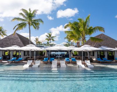 Tropical resort pool with lounge chairs, umbrellas, and palm trees in a serene setting, featuring a restaurant with a thatched roof.