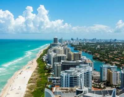 Aerial view of Miami Beach, showcasing turquoise ocean waves, sandy shoreline, and high-rise buildings under a bright blue sky with fluffy clouds.